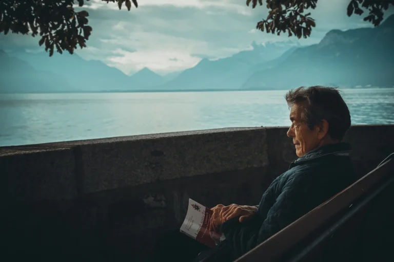 An elderly man, with gray hair and glasses, engrossed in reading a book while seated on a wooden bench.