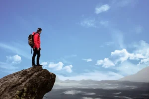 A hiker stands triumphantly atop a mountain peak, carrying a backpack, amidst breathtaking scenery.