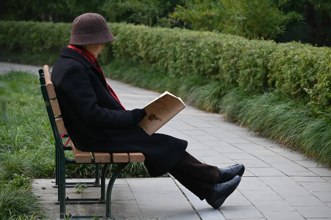 A person engrossed in reading a book while seated on a bench in a serene outdoor setting.