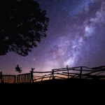 A majestic horse stands before a wooden fence, framed by the beauty and mystery of the Milky Way.