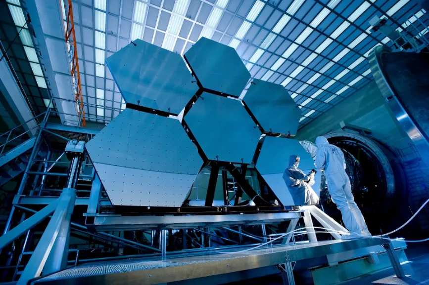 A man in a white suit stands beside a large telescope, possibly the James Webb Space Telescope (JWST) with its impressive specifications.