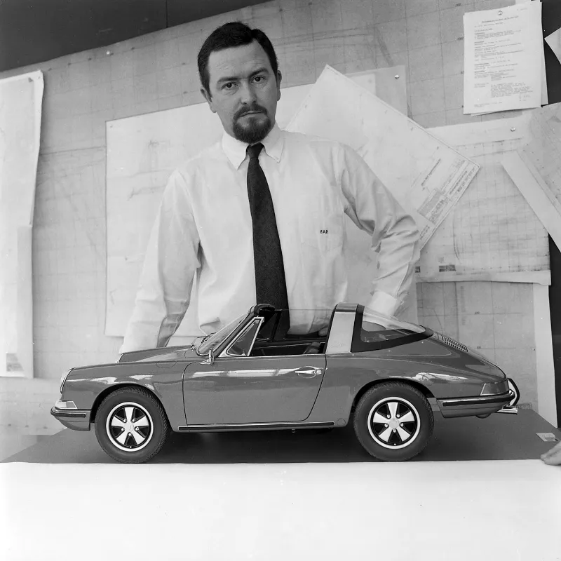 A man in formal attire stands beside a miniature car, showcasing its design and scale.